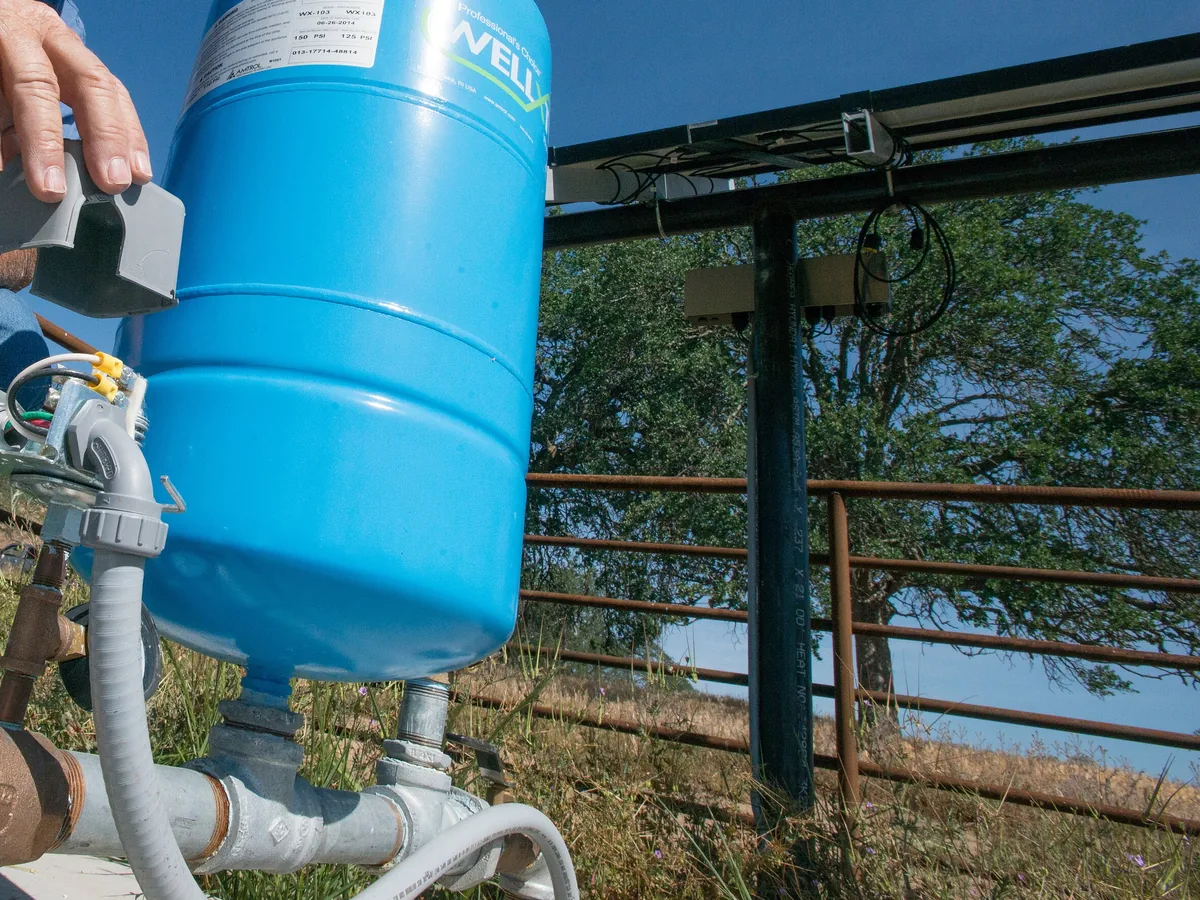 Hand holding the cover of an open pressure switch beside a blue bladder pressure tank
